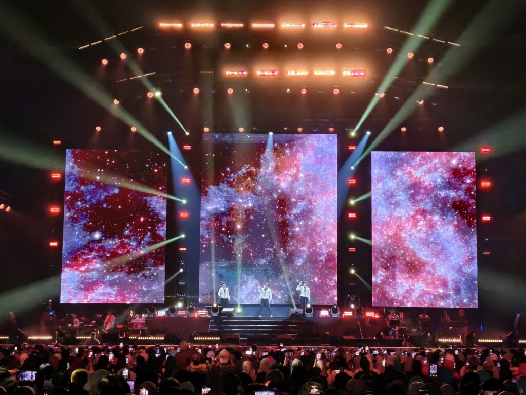 Three performers on stage with large LED screens displaying colorful visuals at a concert.